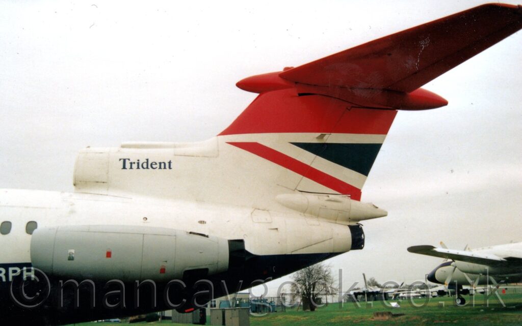 Close-up of the rear fuselage and tail of a 3 engined jet airliner with the engines mounted on the top and sides of the rear fuselage, on display facing to the left outside a museum. The plane is mostly white, with a dark blue belly, with the last 3 letters of the white registration, "RPH" visible below and in front of the side-mounted grey engine pod. The dark blue word "Trident" is on the top of the air intake for the centre engine, mounted on top of the fuselage, in front of the tail. The tail itself is red on the top half and white on the bottom, with a red and blue triangle at the top rear of the white section. Green grass can be seen in the background, under the tail, with a white piston engined airliner on the right edge of the frame, and flat grey sky filling the rest.