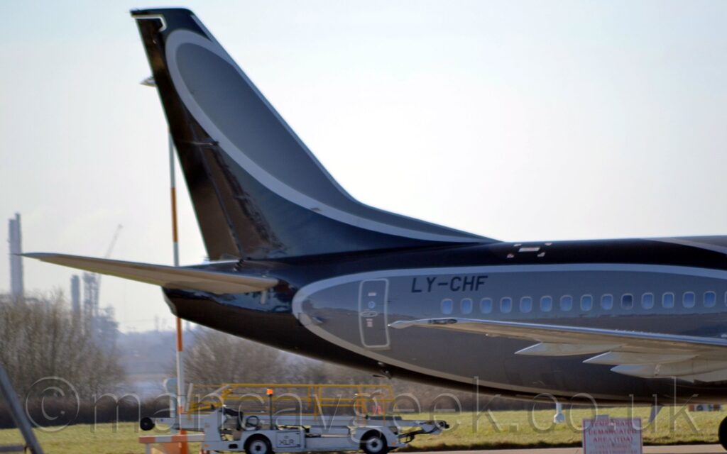 Close-up of the grey and black rear fuselage and tail of a jet airliner parked facing to the right. The body and tail and mostly dark grey, with black swirls outlined in light grey moving across the body, and the black registration "LY-CHF" on the upper rear fuselage. A white mobile baggage loading cart is parked in the middle of the frame in the foreground, with trees visible in the distance, under bright but hazy sky.