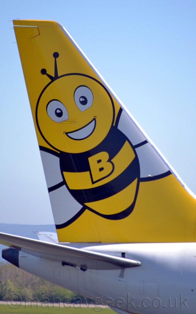 Close-up of the white rear fuselage and bright yellow tail of a passing jet airliner, moving from left to right. The tail has a cartoon-style image of a friendly looking bee with outstretched white wings, and a large uppercase letter "B" in yellow, outlined in black, in the middle of it's torso. Grass and trees in the distance can be seen under the tailcone, with pale blue sky filling the rest of the frame.