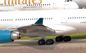 Close-up of the green up-turned wingtip of a twin engined jet airliner taxiing from right to left along a grey taxiway. There are flaps extended from the front and rear of the wing, suggesting it is heading to the runway for takeoff. The plane appears to be almost entirely white, with only the black registration "G-EIDY" to break up the monotony, as well as green engine pods hanging in front of the wings. Grey taxiway fills the foreground, with the white fuselage with gold "Emirates" titles of a larger jet airliner facing to the right filling most of the background, the white fuselage of yet another jet airliner filling the remaining space in the top right corner.
