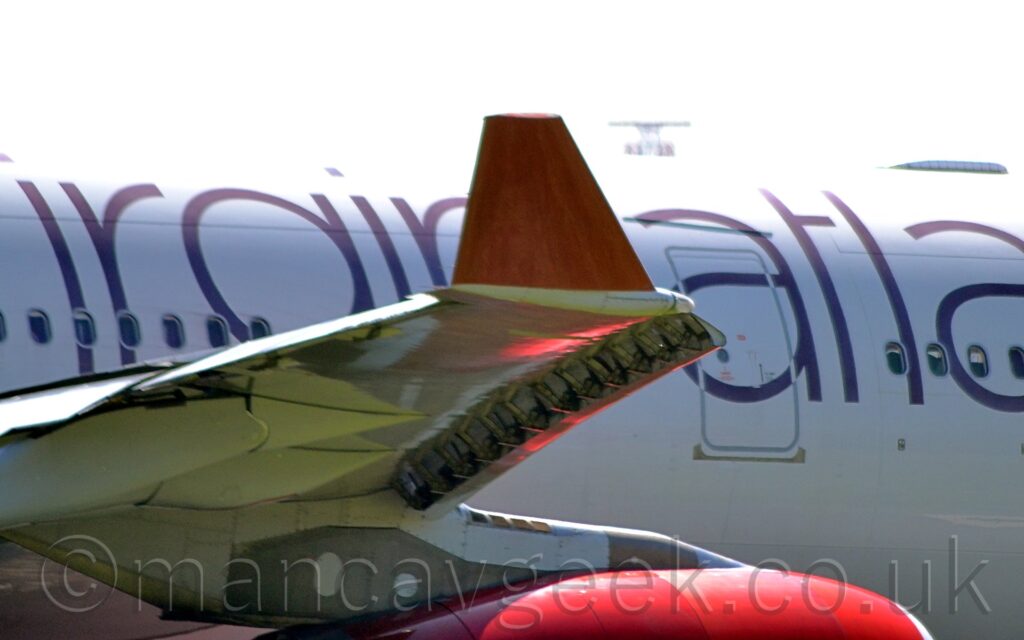 Close-up of the red, up-turned right wingtip of a twin engined jet airliner taxiing from left to right. The plane is mostly white, with purple partial titles "irgin Atla" along the side of the fuselage. A ruby red engine pod hangs under the wing, strongly reflecting sunlight from the top. Flat white sky fills the background.
