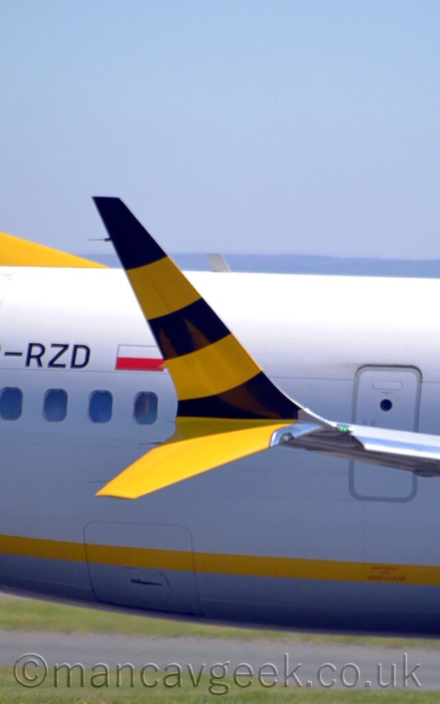 Close-up of the wingtip of a jet airliner taxiing from left to right. The up-turned part of the wingtip has diagonal black and yellow stripes, while the lower, down-turned part is just yellow. In the background, the body of the plane is white, with a thin yellow stripe along the lower fuselage, and the partial black registration "-RZD" next to a small Polish flag on the upper rear fuselage. Green grass lines another taxiway in the background at the bottom of the frame, with blue sky filling the top.