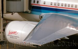 Close-up of the left wingtip and engine of a twin engined jet airliner parked facing to the right at it's gate. The plane is mostly white, with a dark, mid, and light blue stripe running along the body, with a thin red stripe just above that, under the passenger cabin windows. The engine pod hanging under the wing has red, seemingly hand-drawn "Leisure" titles, over black "International Airways" in a block print. A cream coloured airbridge is attached to the fuselage just ahead of the wing.