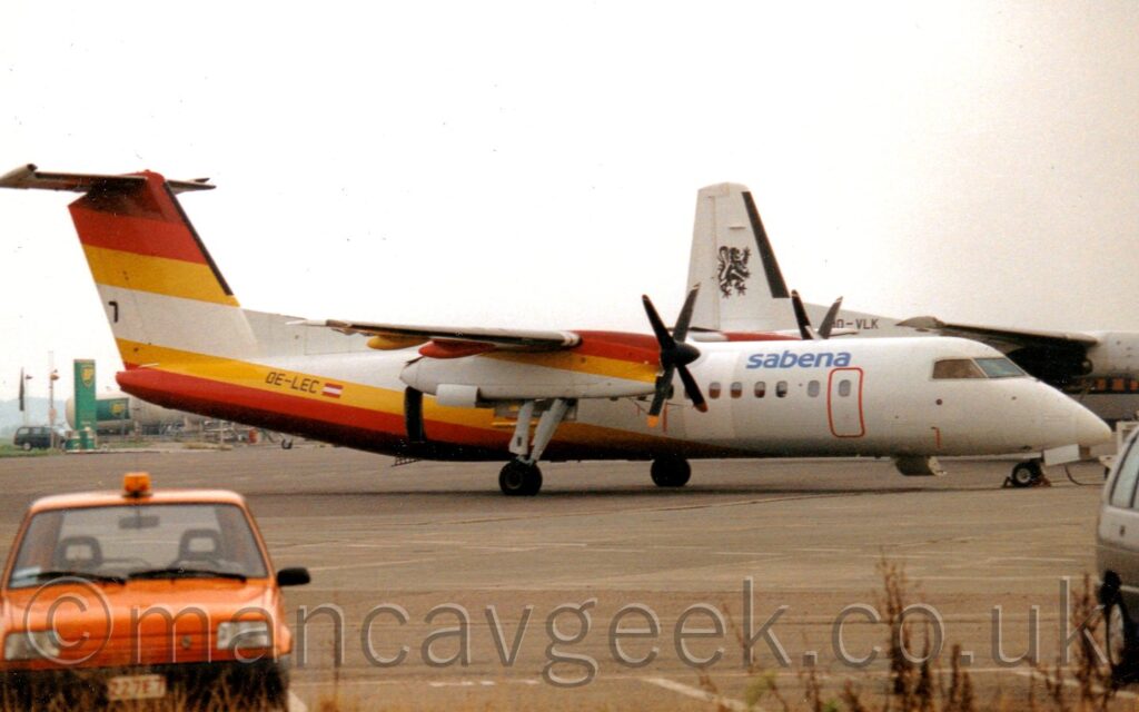 Side view of a high-winged, twin propellor-engined airliner parked facing to the right. The plane is mostly white, with blue "Sabena" titles on the upper forward fuselage, just aft of the forward door outlined in red. There are brown, red, and yellow diagonal stripes on the rear fuselage, appearing in reverse order at the top of the tail and along the top of the engine cowlings over the wings. The black registration "OE-LEC" is on the rear fuselage, next to a small Austrian flag. A small orange car is on the left of the foreground, with an orange light on it's roof. Another plane, this one white with a black rampant lion on the tail, is in the background on the right. Flat white sky fills the rest of the frame.