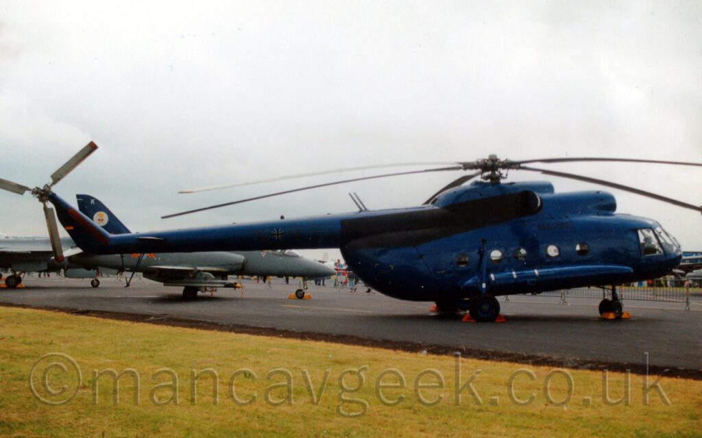 Side view of a fairly large helicopter in an overall deep blue finish, parked on a black taxiway, facing to the right. The black word "Marine" dis painted on the top of the fuselage, below the engine pods and rotor mast, and the black serial "94+12" (the "+" being a stylised black cross outlined in white) in the middle of the long tail boom. A wide grassed section cuts diagonally through the bottom of the frame in the foreground, with a pair of small, dark grey military jets parked in the background on the left. Cloudy grey sky with bright white spots fill the rest of the frame.