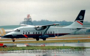 Side view of a rather square, box-sided, high-winged, twin propellor-engined airliner moving from right to left at speed along a black runway, with flaps slightly extended behind the wings, suggesting it is taking off. The plane is light grey on the top half and dark blue on the bottom, with I thin red pin-stripe at the top of the blue, ending in a half arrow-head at the front, just behind the cockpit. There are black "British Airways Express" titles on the upper forward fuselage, the last word being in italics and less bold than the first two words, and the black registration "G-BMAR" is on the upper rear fuselage. The tail is light grey at the bottom, and dark blue at the top, with a dark blue and red triangle on the trailing edge at the top of the grey, and a grey heraldic crest in the middle of the dark blue. Wet black apron and green grass fills the foreground, with more grass in the background leading up to buildings and a metal tower with a white and red radar dish on top in the distance, being swallowed up in grim grey sky.