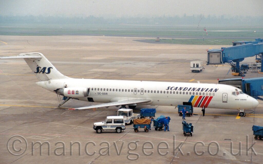 Side view of a twin engined jet airliner, with the engines mounted on the sides of the rear fuselage, parked facing to the right at it's gate. The plane is almost entirely white, with black "Scandinavian" titles on the upper forward fuselage, and diagonal blue, yellow, red, and black bands on the lower forward fuselage. The black registration "SE-DDR" is on the lower rear fuselage, just in front of the white engine pods, which have a series of small squares - red with a white cross, red with a white and black cross, blue with a yellow cross, each with a golden crown on top - on the side, indicating the flags of the 3 Kingdoms this airline is the flag carrier for. A blue airbridge coming from the terminal building on the right of the frame is attached to the forward door on the other side of the plane, while a set of built-in airstairs is deployed from between the engines in the lower rear fuselage. A cargo door is open in the bottom of the body towards the front of the plane, with a blue baggage bin parked in front, and more blue bins hooked up to small white vehicles nearby. Another cargo door is open in the rear fuselage, just below the engine intake, although there are no vehicles near here. Grey concrete apron fills the foreground, with more apron in the background leading up to grass-lined runways in the distance, rapidly being swallowed up in grey mist.