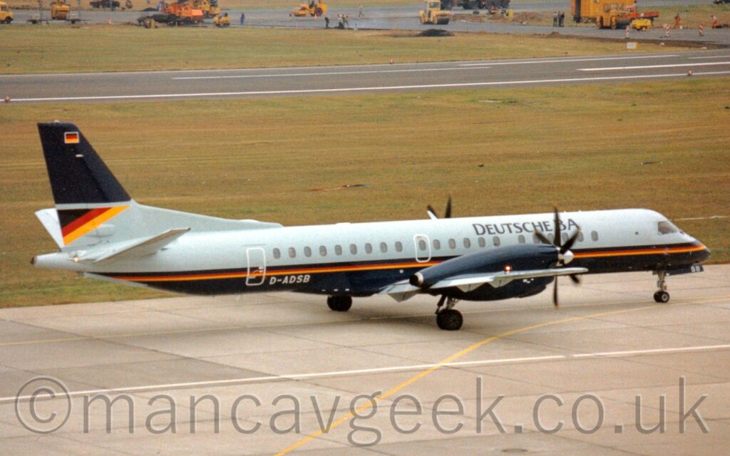 Side view of a long, thin, twin propellor-engined airliner taxiing from left to right along a grey taxiway, it's nose-wheel following a yellow line marked on the surface. The plane has grey upper surfaces, and dark blue lower surfaces, with a black, red, and yellow stripe (the colours of the German flag) running along the body, separating the two colours. There are black "Deutsche BA" titles on the upper forward fuselage, and the white registration "D-ADSB" on the lower rear, just forward of the cabin rear door. The tail is grey and the bottom, with a black, red, and yellow-striped triangle on the trailing edge of the grey, and a dark blue top section, with a small German flag right at the top. Grey concrete apron fills the foreground, with a wide strip of grass separating it from a black runway in the background, with more grass beyond that, and bright yellow road construction vehicles in a newly-constructed patch of black tarmac right at the top of the frame.