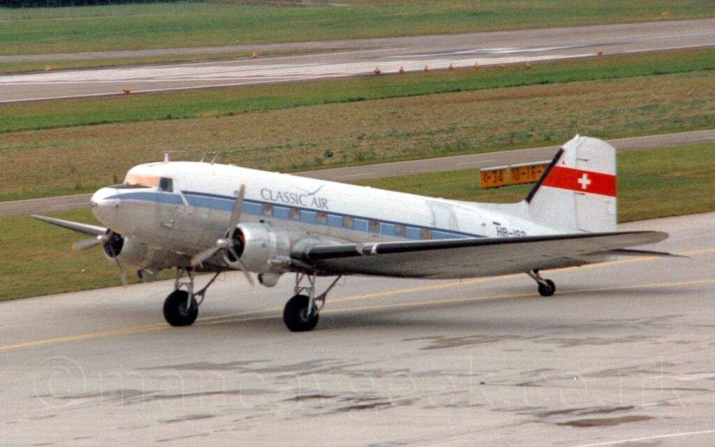 Side view of a twin propellor-engined, World War 2-era airliner taxiing from right to left along a grey concrete taxiway. The plane has a white top half, with a dull bare metal finish on the belly, with a pale blue stripe running along the body, covering the square passenger cabin windows. There are black "Classic Air" titles on the upper forward fuselage, with the black registration "HB-ISB" on the upper rear. The tail is white, with a red band 2/3rds of the way up, containing a white cross. Grey concrete apron fills the foreground, marked with yellow lines for planes to follow, the plane's tail wheel almost precisely on one line that curves off to one side. Green grass fills a large chunk of the background, with a black runway visible in the distance, with more grass on the far side.