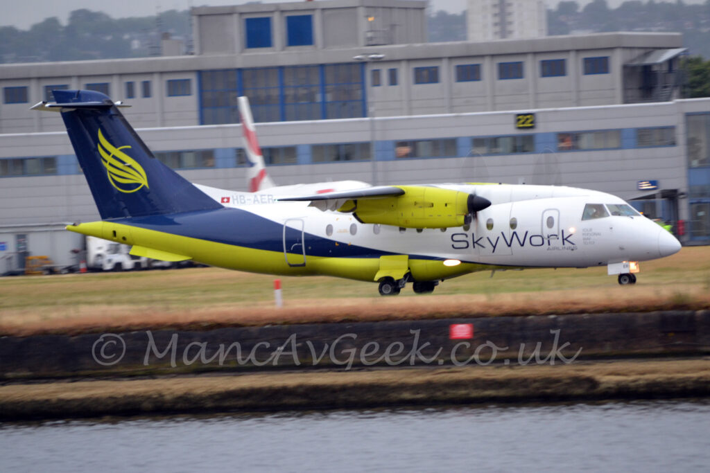 A high-winged, twin propellor-engined airliner moving from left to right along a taxiway hidden by clumps of brown grass. The plane is mostly white on the front and upper surfaces, with dark blue on the rear and upper fuselage, and a lime green, almost fluorescent, belly. There are black "SkyWork" titles on the lower forward fuselage, and the grey registration "HB-AER" on a white fillet at the front of the dark blue tail, next to a small, white, Swiss flag. Bright lime-green feathers over the lime green outline of a circle are on the tail, with similarly bright green engine pods hanging under the wings. A body of water stretches across the foreground, a couple of metres lower than the ground. Grass in the background leads up to a grey building that stretches across the frame, with grey sky beyond that.