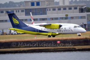 A high-winged, twin propellor-engined airliner moving from left to right along a taxiway hidden by clumps of brown grass. The plane is mostly white on the front and upper surfaces, with dark blue on the rear and upper fuselage, and a lime green, almost fluorescent, belly. There are black "SkyWork" titles on the lower forward fuselage, and the grey registration "HB-AER" on a white fillet at the front of the dark blue tail, next to a small, white, Swiss flag. Bright lime-green feathers over the lime green outline of a circle are on the tail, with similarly bright green engine pods hanging under the wings. A body of water stretches across the foreground, a couple of metres lower than the ground. Grass in the background leads up to a grey building that stretches across the frame, with grey sky beyond that.