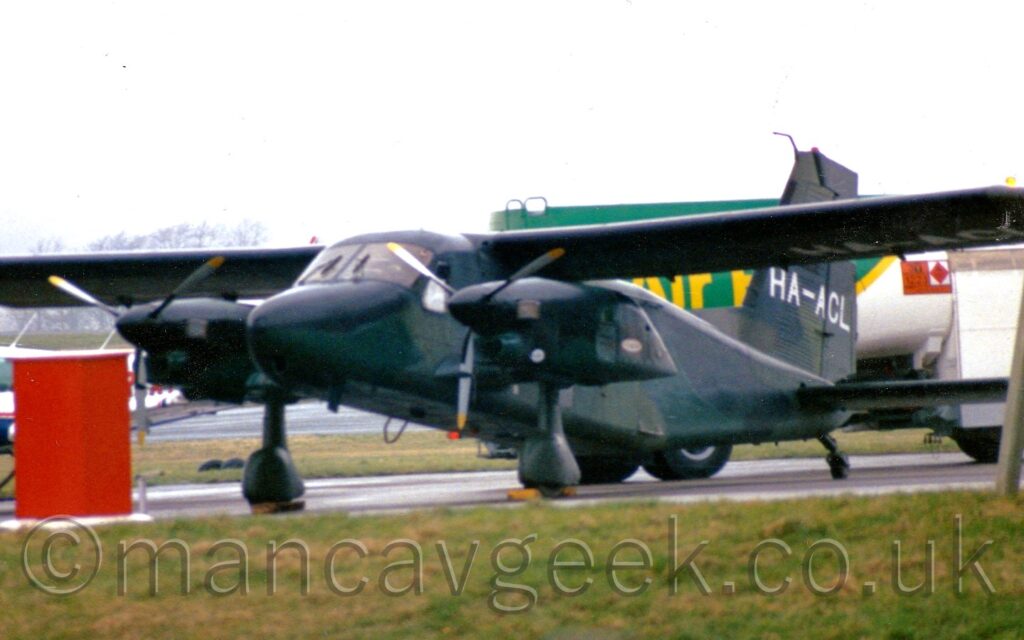 Front side view of a high-winged, twin propellor-engined light aircraft, parked facing to the left on a grey taxiway, with the engines mounted on stub wings attached to the bottom of the fuselage, below the main wings. The plane is covered in a camouflage scheme made up from various shades of green, with the white registration "HA-ACL" on the tail. Green grass fills the foreground, with a tall, red metal box in front of the planes right wing. A green and white fuel truck is in the background, under flat white sky.