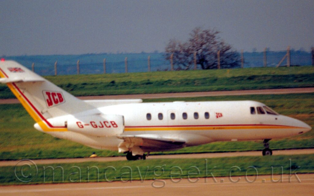 Side view of a twin engined BizJet taxiing from left to right. The plane is mostly white, with a thin red and slightly thicker white stripe running along the body, sweeping up along the trailing edge of the tail. The red registration "G-GJCB" is on the side of the white engine pods mounted on the sides of the rear fuselage. There is a red "JCB" logo inside the thin outline of an off-angle quadrilateral, a design repeated in a smaller form just in front of the front windows in the passenger cabin. A small British flag is painted right at the tip of the tail. A strip of green grass separates a taxiway at the front of the frame with the taxiway the plane is on, with more grass in the background leading up to a runway, and a chain-link fence on concrete columns at the top of a slight grassy rise, with tree-covered hills in the distance being swallowed up in hazy grey sky.