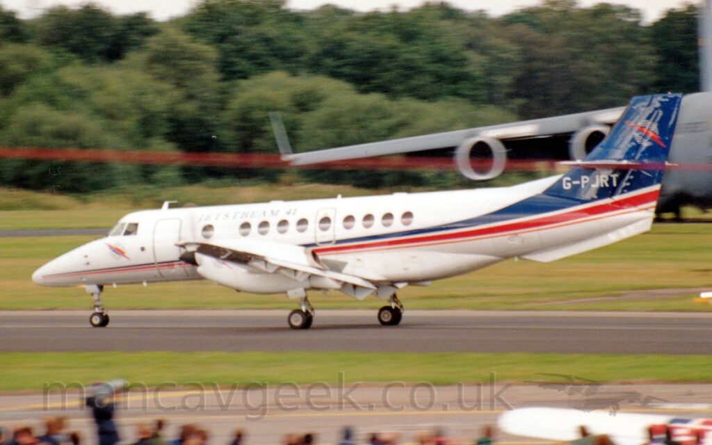Side view of a twin propellor-engined airliner moving at speed along a black runway, with flaps hanging down from the rear of the wings, suggesting it has just landed. The plane is white, with a thin red, blue, and orange stripe running along the lower body, the blue and red getting wider as it moves rearwards, until the blue fills the tail. There are black "Jetstream 41" titles on the upper forward fuselage, and the white registration "G-PJRT" at the base of the tail, under the horizontal stabiliser. The heads of people in the crowd line a grey taxiway in the foreground, separated from the runway by a strip of green grass. More grass in the background leads up to trees in the distance, with the body of a grey, 4 engined military jet transport on the left of the background, , it's wing hanging out over most of the frame with engine pods below.