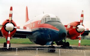 Front-side view of a twin propellor-engined military transport trainer aircraft parked facing to the right on tarmac behind a low, white, metal fence. The plane has a large red stripe running along the middle of the body, covering the passenger cabin windows, with a red roof and dark blue belly,. The engines mounted on the wing are the same bright red, as is the tail. The nose-cone, and top of the nose in front of the cockpit, is painted matt black. Green grass can be seen in the background, under the plane, with flat grey sky filling the rest of the frame.