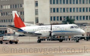 Side view of a twin propellor-engined airliner parked facing to the right. The plane is mostly white, with a thin light and dark blue stripe running along the body, and thin, black, seemingly hand-written "Tatra" titles on the upper fuselage. The tail has what seems to be a large red mountain with white sky at the top. A large expanse of apron fills the foreground, with baggage carts scattered in the background, and a pair of grey office blocks filling the frame in the distance.