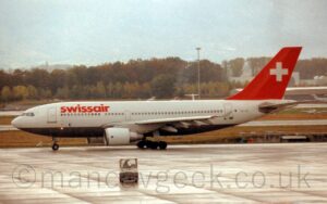 Side view of a twin engined jet airliner taxiing from right to left. The plane is largely white, with a grey belly, and red "SwissAir" titles on the upper forward fuselage, and the black registration "HB-IPI" on the lower rear. The tail is red, with a large white cross in the middle. A large grey concrete apron area with a small truck in the middle fills the foreground, with grass and a runway in the background leading up to trees in the distance on the left, and a grey and blue hangar with a couple of smaller planes parked outside on the right. Tree covered hills beyond that are rapidly swallowed up in misty cloud.
