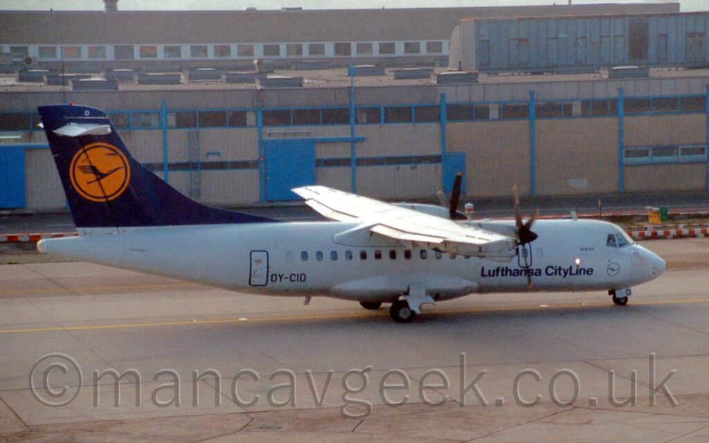 Side view of a high-winged, twin propellor-engined airliner taxiing from left to right along a grey taxiway, following a yellow line marked on the surface. The plane has a white body, with dark blue "Lufthansa CityLine" titles on the lower forward fuselage, and the black registration "OY-CID" on the lower rear, just forward of the rear cabin door. The tail is dark blue, with a bright yellow circle in the middle, containing a highly stylised blue flying bird. Grey concrete taxiway fills the foreground, with a sandy-brown building stretches across the frame in the background.
