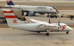 Side view of a high-winged, twin propellor-engined airliner taxiing from left to right, following a yellow line marked on a grey taxiway. The plane is mostly white, with a grey belly, and small "Austrian" titles on the lower forward fuselage, partially obscured by the undercarriage legs coming down from the engine pods under the wings. There is a stylised, red arrowhead on the forward fuselage, behind the cockpit, and the black registration on a white fillet at the front of the tail. The white tail has a pair of thick red lines, representing the Austrian flag, at the top, and a stylised, red, upper-case letter "R" at the bottom, the vertical bar replaces with another red arrowhead, this one pointing rearwards. Grey concrete apron fills the foreground, with a sandy-brown building in the background on the left, and a white, twin engined jet airliner with a blue and yellow tail in the middle, as well as low, lime green tug on the right.