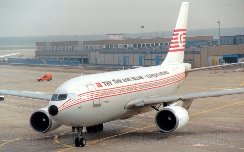 Three-Quarter left front view of a twin engined jet airliner taxiing from right to left, heading for the bottom left corner of the frame. The plane has a white top half and grey lower half, with thin red pinstripes running along the body, covering the passenger cabin windows. There is large black text "THY" on the upper forward fuselage, immediately aft of a small, red and white Turkish flag, with smaller "Turk Hava Yollari - Turkish Airlines" text behind that, spreading over most of the rest of the upper fuselage. The tail is white, with 6 red stripes running through the middle, overlaid with a white circle containing a highly stylised red flying bird. Grey concrete apron fills the foreground and most of the background, with a pair of sandy-brown buildings stretching across the frame from the right, under hazy grey sky.