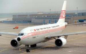 Three-Quarter left front view of a twin engined jet airliner taxiing from right to left, heading for the bottom left corner of the frame. The plane has a white top half and grey lower half, with thin red pinstripes running along the body, covering the passenger cabin windows. There is large black text "THY" on the upper forward fuselage, immediately aft of a small, red and white Turkish flag, with smaller "Turk Hava Yollari - Turkish Airlines" text behind that, spreading over most of the rest of the upper fuselage. The tail is white, with 6 red stripes running through the middle, overlaid with a white circle containing a highly stylised red flying bird. Grey concrete apron fills the foreground and most of the background, with a pair of sandy-brown buildings stretching across the frame from the right, under hazy grey sky.