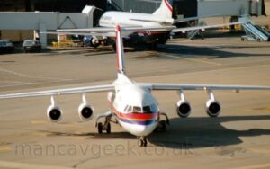Forward view of a high-winged, 4 engined jet airliner taxiing from left to right, almost directly towards the camera. The plane is white, with a red and purple stripe with yellow piping in the middle running along the body, and a red tail with a purple stripe with yellow piping at the bottom. It's nose wheel is turned to it's left, as it turns to the right of the frame. In the background, a grey and dark blue, twin engined jet airliner facing away from the camera, has an airbridge connected to it's forward fuselage, the other end of the bridge connected to a grey buildin