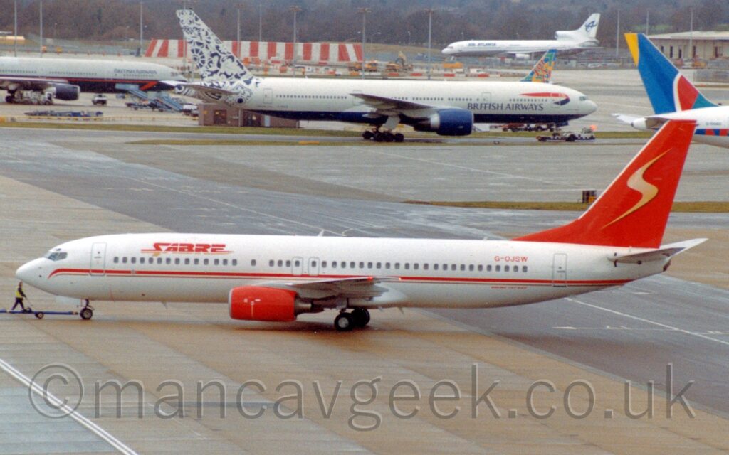 Side view of a twin engined jet airliner , facing to the left, but being pushed back to the right by an out-of-frame tug attached to the nose-wheel by a long blue pole. There are red "Sabre" titles with an orange drop shadow on the upper forward fuselage, and the red registration "G-OJSW" on the upper rear. There is a red and gold stripe running along the body, between the passenger cabin windows and the wing, , and red engine pods mounted under the wings. The tail is the same bright red, with a highly-stylised golden letter "s" in the middle. Grey concrete squares make up a large apron area that fills the foreground and much of the background, with larger twin engined jet airliners facing to the right in the background, with a 3 engined airliner in the distance facing to the left, with trees running across the back of the frame.