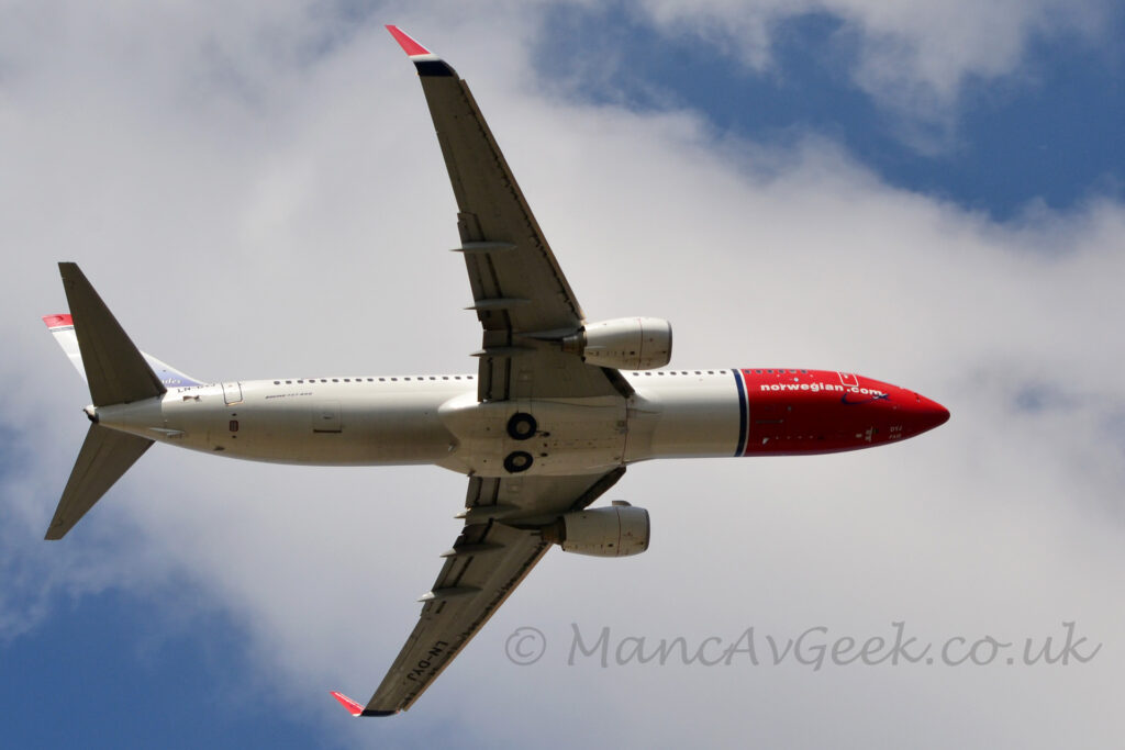 View from beneath of a twin engined jet airliner flying from left to right at a low altitude, with undercarriage tucked away and flaps extended from the rear of the wings, with the nose raised, suggesting it has just taken off. The body is mostly white, with a red nose and forward fuselage, separated from the rest of the body by a thin blue band around the body., with white "NorwegianDotCom" titles on the lower forward fuselage. The black registration "LN-DYJ" is on the upper rear fuselage, just in front of the horizontal stabiliser, and repeated on the underside of the outer panels on the left wing. The up-turned wing-tips are the same bright red as the forward fuselage, while the tail is a purpley-blue at the bottom, most of the rest is white, and a thick red and thin blue stripe at the top. Fluffs of white cloud stretch from the top left corner to the bottom right, with blue filling the rest of the background.