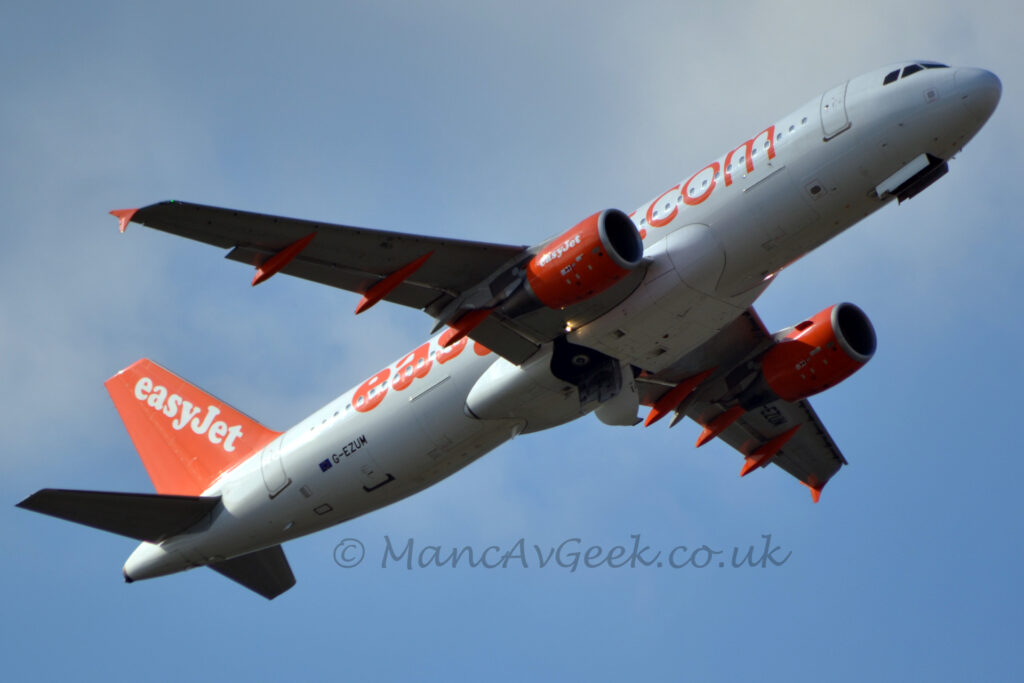 Low side view of a twin engined jet airliner flying from left to right at a very low altitude, in the middle of tucking it's undercarriage away, with flaps extended from the rear of the wings, and nose raised, suggesting it has just taken off. The plane's fuselage is almost entirely white, with large, orange, billboard-style "EasyJetDotCom" titles along the side, partially obscured by the right wing. The black registration "G-EZUM" is on the lower rear fuselage, just ahead of a small EU flag, the registration repeated in a larger form under the left #wing. The bright orange engine pods hanging under the wings have white text "EasyJet" on the sides. The flap-track covers under the wings are also bright orange. The tail is the same bright orange, with large, white, diagonal "EasyJet" titles running along the leading edge. Pale blue sky fills the background, with wisps of fluffy white cloud in the top right corner and the middle of the left edge of the frame.
