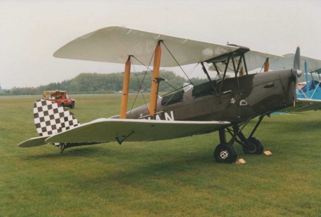 Side view of a single-engined biplane with a pair of open cockpits, parked facing to the right on grass. The plane's body is a dark brown, with the registration painted on the side in white, behind the wings - only the last 2 letters, "AN" can be made out. The tail is covered in a brown and white checkerboard pattern. The wings are white, with varnished brown struts 2/3rds of the way out from the body supporting the upper wings. Green grass fills the foreground, with a blue biplane in the background on the right, and more grass filling the rest of the background, with trees in the distance under grey sky.