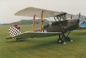 Side view of a single-engined biplane with a pair of open cockpits, parked facing to the right on grass. The plane's body is a dark brown, with the registration painted on the side in white, behind the wings - only the last 2 letters, "AN" can be made out. The tail is covered in a brown and white checkerboard pattern. The wings are white, with varnished brown struts 2/3rds of the way out from the body supporting the upper wings. Green grass fills the foreground, with a blue biplane in the background on the right, and more grass filling the rest of the background, with trees in the distance under grey sky.