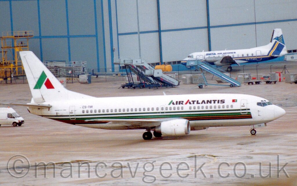 Side view of a twin engined jet airliner taxiing from left to right, across a large area of grey concrete apron. The plane is mostly white, with black "Air Atlantis" titles on the upper forward fuselage, and the black registration "CS-TIR" on the upper rear. A series of dark green pin-stripes run along the lower fuselage, the gaps in the lower ones filled in with red, orange, and light green. The tail is white, with a large, stylised upper-case letter "A" with the left side being light green, and the right side being red. A white van is following the plane on the left of the frame. Grey concrete apron fills the foreground, with more apron leading up to a large grey hangar with blue piping in the background. A smaller, blue and white, twin propellor-engined airliner is parked on the left of the frame, in front of the hangar.