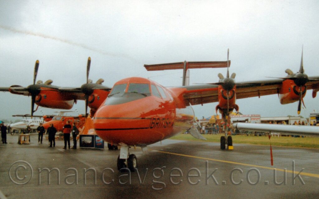 Low, almost head-on view of a high-winged, 4 propellor-engined airliner parked on damp black tarmac, facing to the left. The plane is almost entirely a bright orange, with a air of thin black lines running along the body, the word "British" between them under the cockpit windows. A built-in set of stairs hangs down from the side of the rear fuselage. A smaller, twin propellor-engined airliner in similar colours is visible in the left of the background under this one's wing, with a group of people standing around it, with another, smaller again twin engined white BizProp beyond that. A damp grey taxiway fills the foreground, as well as a good bit of the left of the background, with white and grey sky filling the top of the frame.
