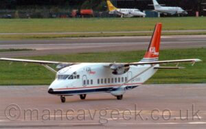 Front side view of a rather boxy-looking, high-winged, twin propellor-engined airliner taxiing from right to left. The plane is mostly white, with a thin red pinstripe and a thicker blue stripe running along the bottom of the body, and the black registration "G-DASI" on the side of the rear fuselage. There are a couple of words printed on the side of the upper forward fuselage - "Gill", in blue, with "Airways" in red pretty much underneath it, looking slightly hand drawn. The tail has a couple of thin dark blue stripes on the bottom, with the rest being a flat red, with the white words "Gill Airways" towards the middle. Grey apron fills the foreground, with a couple of large grassed areas and a wide, black runway filling most of the background, with a pair of small, white, twin propellor-engined airliners parked against trees on the far side.