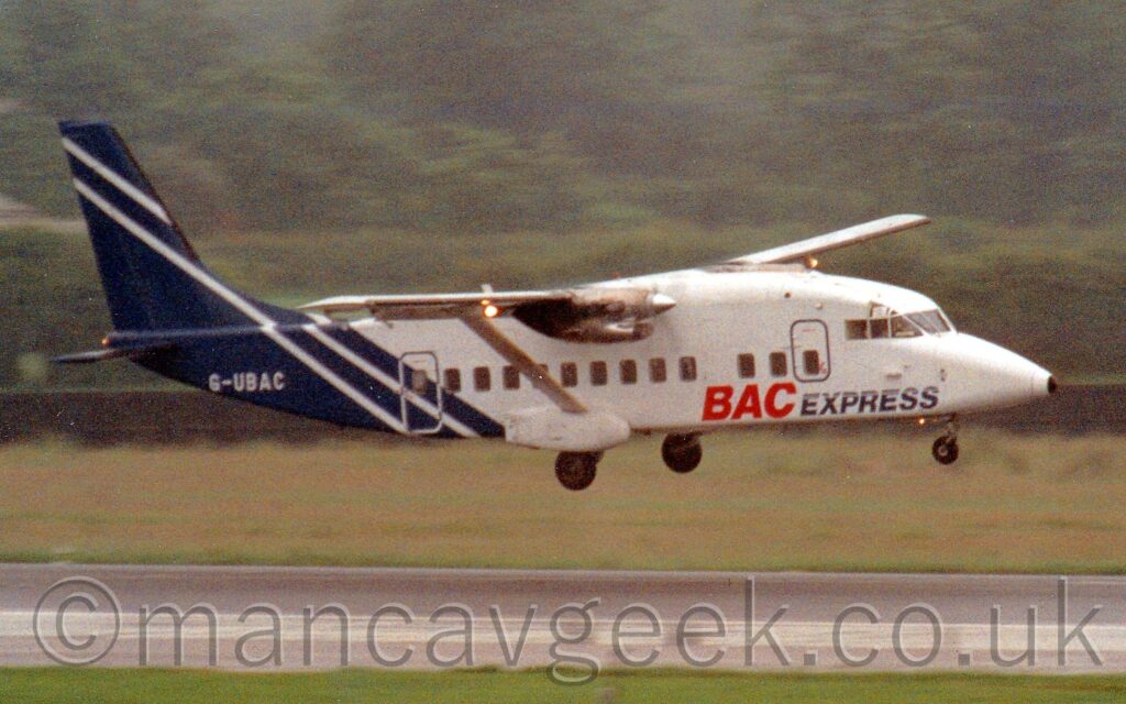 Side view of a very square, high-winged, twin propellor-engined airliner flying from left to right at a very low altitude, with undercarriage lowered and flaps extended from the rear of the wings, suggesting it is about to land. The plane is mostly white, with a dark blue rear fuselage and tail. There are red and blue "BAC Express" titles on the sides of the lower forward fuselage, and the white registration "G-UBAC" on the side of the rear fuselage. Green grass and black runway fill the foreground, with more grass in the background leading up to bushes and trees in the distance.