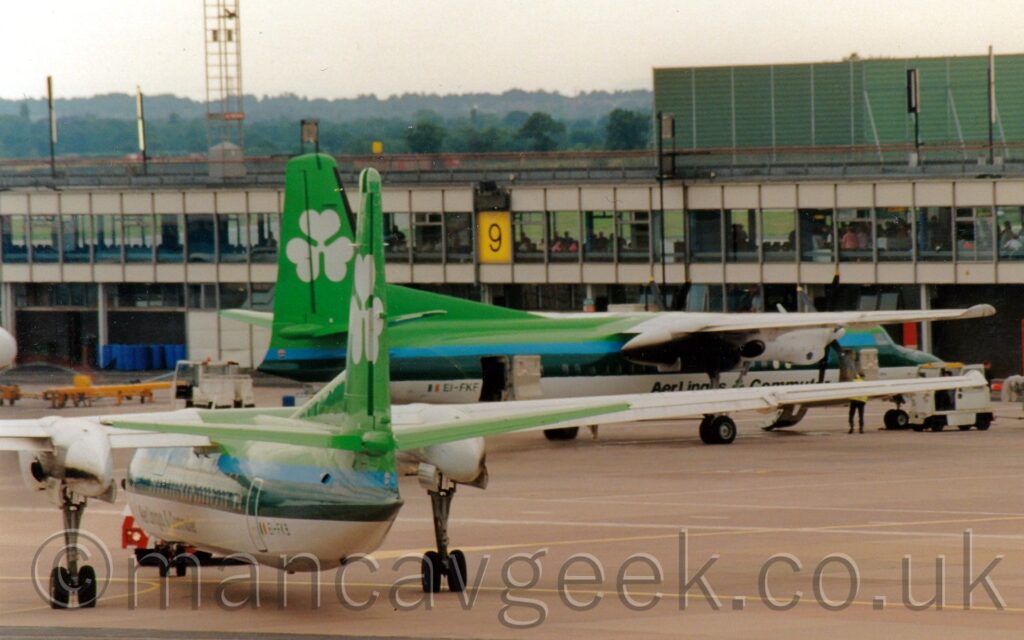 Rear view of a high-winged, twin propellor-engined airliner facing to the left but being pushed back to the right by a red and white tug that is almost hidden by the plane. The plane is a bright green on top, with a blue stripe below that, and a thick dark green stripe running along the body, covering the passenger cabin windows. The lower half is white, with a grey belly. There are dark green "Aer Lingus Commuter" titles on the lower forward fuselage, and the dark green registration "EI-FKB" next to a small Irish flag on the lower rear. A near identical airliner is parked facing to the right in the background, nose-in to a heavily glazed terminal building that stretches across the screen, the biggest difference being that this one has the registration "EI-FKF" at the rear. Grey concrete apron fills the foreground, with trees meeting bright but hazy sky in the distance.