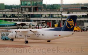 Rear side view of a high-winged, twin propellor-engined airliner facing to the left but behind pushed back to the right by a low blue tug attached to the planes nosewheel by a long blue pole. The plane is mostly white, with black "Lufthansa Cityline" titles on the lower forward fuselage, partially obscured by the undercarriage legs coming down from the rear of the engine pod on the left wing, and the black registration "D-BELT" on the side of the rear fuselage, next to a small German flag. The tail is dark blue, with the outline of a yellow circle and a highly stylised image of a flying bird, made up of 5 yellow lines. Grey concrete apron fills the foreground, with a terminal building stretching all the way across the background, with a green and white, high-winged, propellor-engined airliner parked nose-in. In the distance, there are a grey hangar on the right, and trees in the centre and on the left, under bright but hazy sky.