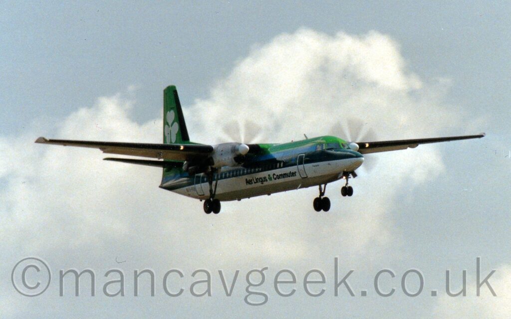 Forward side view of a high-winged, twin propellor-engined airliner flying from left to right at a low altitude, with undercarriage lowered and flaps extended from the rear of the wings, suggesting it is about to land. The upper fuselage is bright green, with a blue stripe below that, a dark green stripe covering the passenger cabin windows, and a thick white stripe above a grey belly. There are dark green "Aer Lingus Commuter" titles on the lower forward fuselage, with a bright green shamrock in the middle, and the registration "EI-FKE" next to a small Irish flag on the lower rear. A large white shamrock fills the bright green tail. Pale blue sky with large fluffs of white cloud fill the rest of the frame.