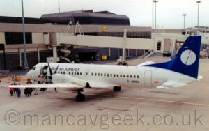 Rear side view of a twin propellor-engined airliner parked facing to the left, nose-in to a terminal building. The plane is predominantly white, with black "British Airways" titles on the upper forward fuselage, and the registration "G-BRLY" on the lower rear. The tail is blue, with a large white circle in the middle. The door in the front of the passenger cabin is open, with passengers queuing on the apron to climb the built-in airstairs to get onboard. Grey concrete apron fille the foreground, with a grey and black terminal building with airbridges hanging off the side filling most of the background, under grey sky.