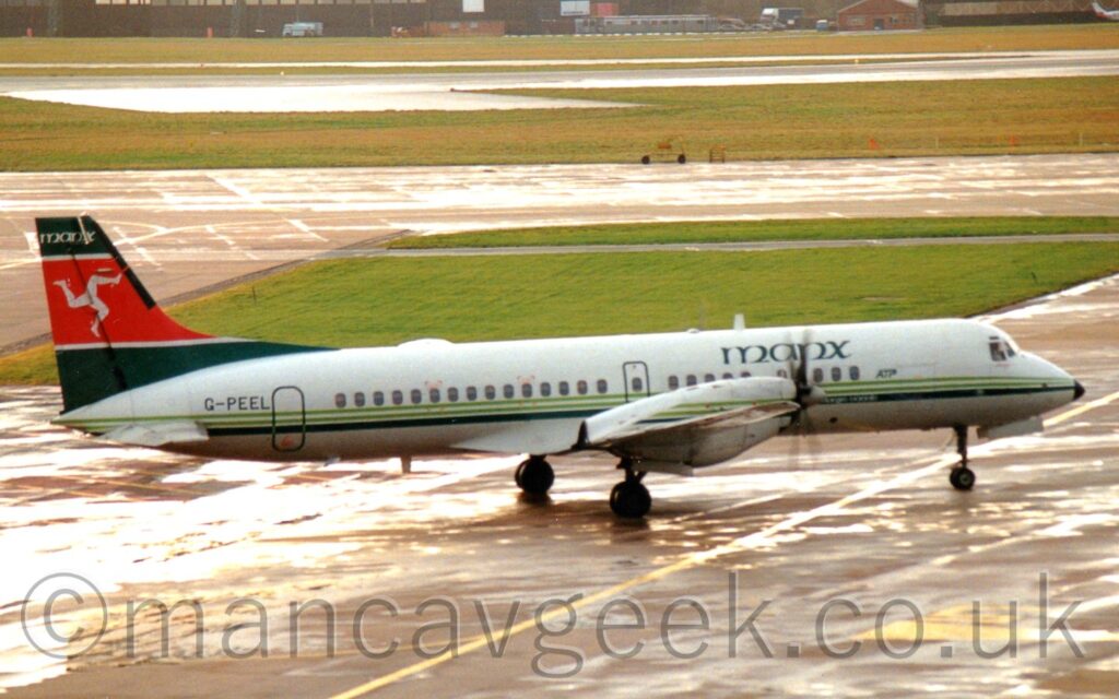 Side view of a twin propellor-engined airliner taxiing from left to right and turning slightly to it's left, across damp grey apron marked with yellow lines. The plane is mostly white, with 2 thin, light green stripes and a thicker dark green stripe running along the body, and green "Manx" titles on the upper forward fuselage., and the dark green registration "G-PEEL" on the side of the rear fuselage. The tail is dark green, with a thick red stripe through the middle outlined in white, and with a white tri-skelion (3 armoured legs running in a circle, joined at the hip) in the middle. Sunlight shines on puddles on grey concrete apron in both the foreground and background, with a large parge of grass in the background leading up to a similarly damp taxiway, with more grass leading up to a black runway lined with grass in the distance.