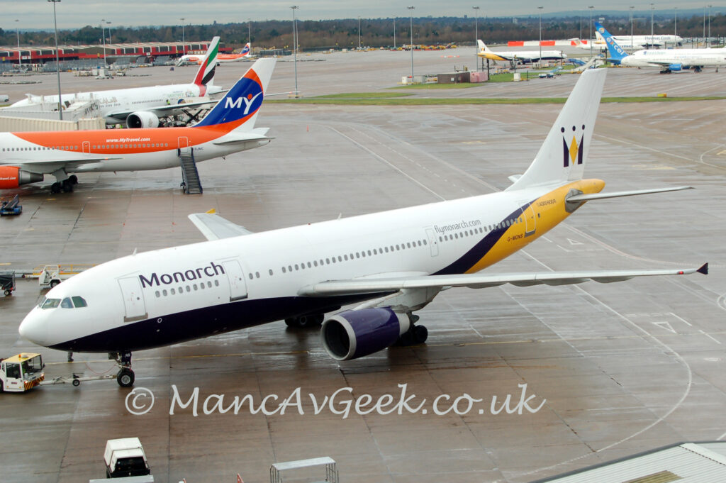 Side view of a twin engined jet airliner facing to the left but being pushed back to the right by a very low, white tug attached to the planes nose wheel by a long white pole. The plane is mostly white, with a dark purple belly, and a yellow rear fuselage. There are black "Monarch" titles on the upper forward fuselage, and the black registration "G-MONS" on the yellow lower rear fuselage. There is additional purple text "FlyMonarchDotCom" on the upper rear fuselage. Slightly damp grey concrete apron fills the foreground, with more apron filling a large chunk of the background, and scattered jet airliners , mostly nearby in the top left corner, and further out in the top right. trees at the top of the frame mark the extent of the airfield in that direction.