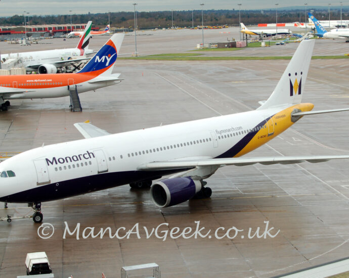Side view of a twin engined jet airliner facing to the left but being pushed back to the right by a very low, white tug attached to the planes nose wheel by a long white pole. The plane is mostly white, with a dark purple belly, and a yellow rear fuselage. There are black "Monarch" titles on the upper forward fuselage, and the black registration "G-MONS" on the yellow lower rear fuselage. There is additional purple text "FlyMonarchDotCom" on the upper rear fuselage. Slightly damp grey concrete apron fills the foreground, with more apron filling a large chunk of the background, and scattered jet airliners , mostly nearby in the top left corner, and further out in the top right. trees at the top of the frame mark the extent of the airfield in that direction.