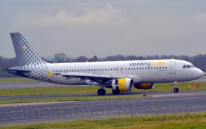 Side view of a twin engined jet airliner taxiing from left to right along a black taxiway. The plane is mostly white, with grey, lowercase "vueling" and yellow "com" titles on the upper forward fuselage. The tail and rear fuselage start off with small grey dots towards the front, the dots gradually getting larger and merging into a solid wall of grey, with a single yellow dot up towards the top. Green grass lines the sides of a pair of black taxiways in the foreground, and more in the background separates the plane from a black runway, with trees in the distance meeting grey sky.