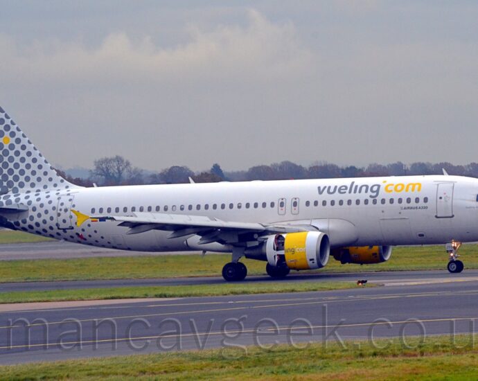 Side view of a twin engined jet airliner taxiing from left to right along a black taxiway. The plane is mostly white, with grey, lowercase "vueling" and yellow "com" titles on the upper forward fuselage. The tail and rear fuselage start off with small grey dots towards the front, the dots gradually getting larger and merging into a solid wall of grey, with a single yellow dot up towards the top. Green grass lines the sides of a pair of black taxiways in the foreground, and more in the background separates the plane from a black runway, with trees in the distance meeting grey sky.