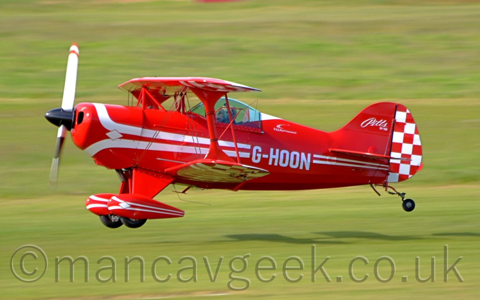 Side View of a single engined, single seat bi-plane light aircraft flying from right to left at a very low latitude, less than a metre above a grass runway. The plane is mostly red, with a pair of white stripes running along the body, overlaid with the white registration "G-HOON", with similar white stripes running the length of the spats on the main wheels, under the wings. The front of the tail is red, with white "Pitts S-1S"text, while the rear is covered in a red and white checkerboard pattern, which can also be seen on the outer panels of the under side of the wings. A person wearing a white flying helmet can be seen in the single seat cockpit, under a domed canopy. Grrn grass fills the rest of the frame, with the plane casting a strong shadow on the ground behind it, suggesting that the sun is coming from the top left corner.