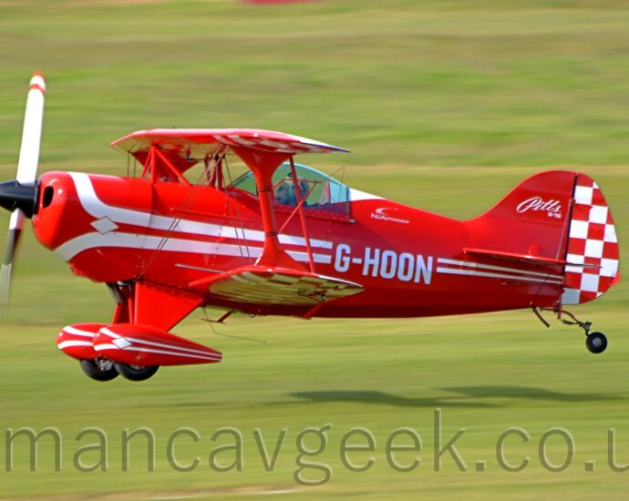Side View of a single engined, single seat bi-plane light aircraft flying from right to left at a very low latitude, less than a metre above a grass runway. The plane is mostly red, with a pair of white stripes running along the body, overlaid with the white registration "G-HOON", with similar white stripes running the length of the spats on the main wheels, under the wings. The front of the tail is red, with white "Pitts S-1S"text, while the rear is covered in a red and white checkerboard pattern, which can also be seen on the outer panels of the under side of the wings. A person wearing a white flying helmet can be seen in the single seat cockpit, under a domed canopy. Grrn grass fills the rest of the frame, with the plane casting a strong shadow on the ground behind it, suggesting that the sun is coming from the top left corner.