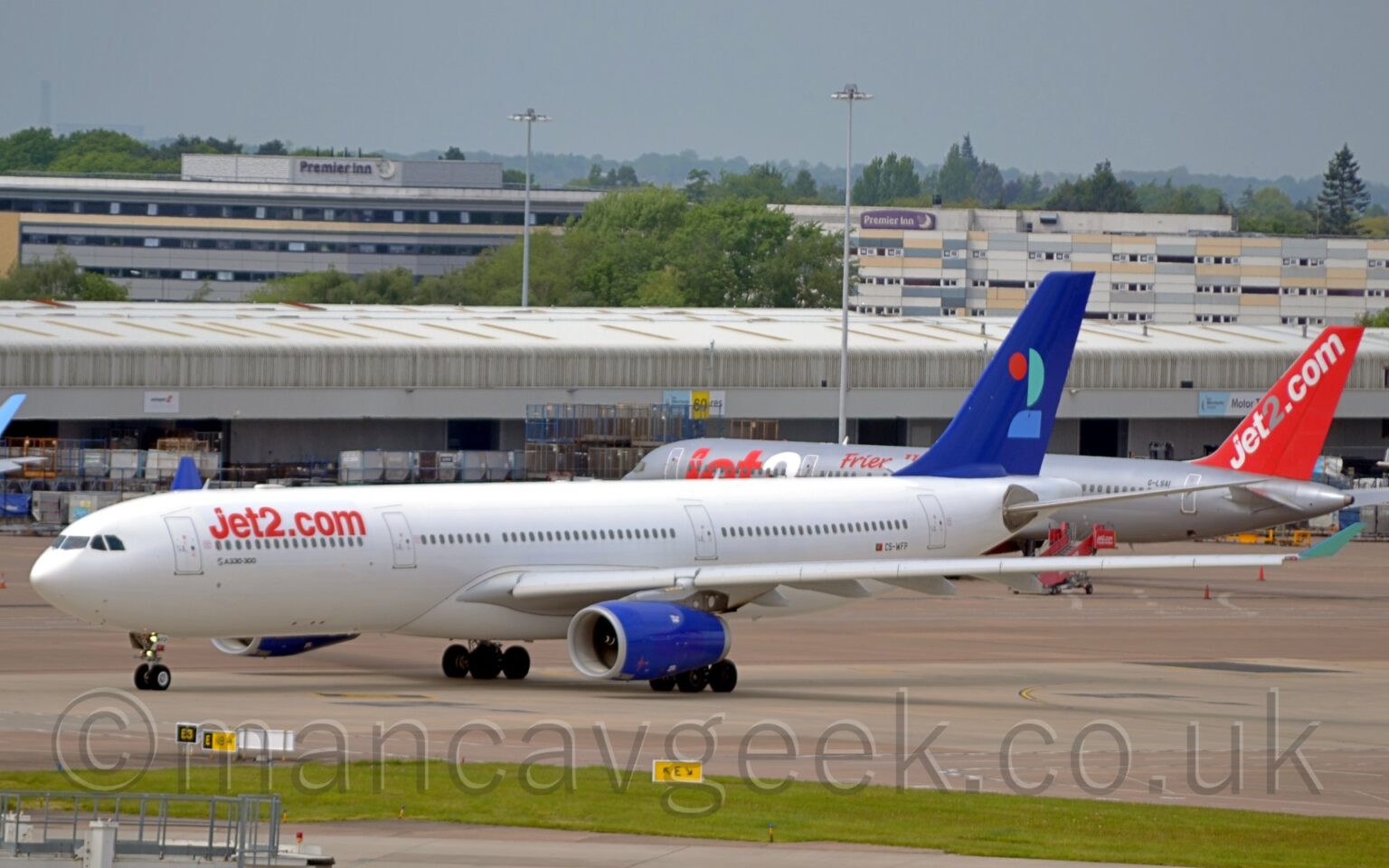 Side view of a white, twin engined jet airliner taxiing from right to left, along a grey taxiway. The plane has red "Jet2DotCom" titles on the upper forward fuselage, and the black registration "CS-WFP" just aft of a small Portugese flag on the lower rear. The tail is a deep blue, with a stylised number "2" made up of red, mint green, and light blue sections. The engine pods mounted under the wings are the same deep blue as the tail, with the up-turned wing-tips are the same mint green as in the number on the tail. A strip of green grass stretches across the foreground, with a grey cargo building stretching across the background with a grey and red jet airliner parked in front on the right. A couple of grey buildings with hotel logos on top are visible over the top off that building, with trees beyond them, under hazy grey skies.