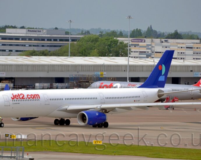 Side view of a white, twin engined jet airliner taxiing from right to left, along a grey taxiway. The plane has red "Jet2DotCom" titles on the upper forward fuselage, and the black registration "CS-WFP" just aft of a small Portugese flag on the lower rear. The tail is a deep blue, with a stylised number "2" made up of red, mint green, and light blue sections. The engine pods mounted under the wings are the same deep blue as the tail, with the up-turned wing-tips are the same mint green as in the number on the tail. A strip of green grass stretches across the foreground, with a grey cargo building stretching across the background with a grey and red jet airliner parked in front on the right. A couple of grey buildings with hotel logos on top are visible over the top off that building, with trees beyond them, under hazy grey skies.