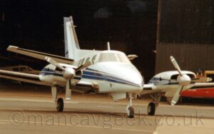 Front view of a twin propellor-engined BizProp parked facing slightly to the right. The plane is white, with a thick dark blue stripe running along the side of the fuselage, as well as along the sides of the engine pods mounted in front of the wings. Grey concrete apron fills the foreground, with a large black hangar filling the background, some of it's inhabitants visible through the large, open doors.