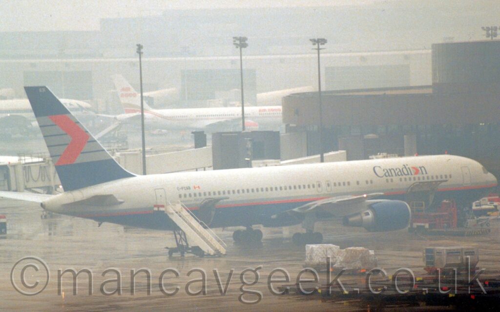 A twin engined jet airliner parked facing to the right, nose-in to a brown and black terminal building he plane is light grey on the upper half, the bottom half being a dark blue, with a thin dark grey and red stripe separating the 2 colours. There are large, dark blue "Canadian" titles on the upper forward, the last letter "a" replaced with a red chevron pointing to the right so it can easily be replaced with an "e" in French-speaking parts of Canada. There is a cargo door hinged open in the lower forward fuselage, with a red cargo loader in the lifted position beside it. A smaller cargo door is hinged upwards in the lower rear, next to a white set of mobile airstairs parked by the open rear cabin door. The black registration "C-FCAB" is on the upper rear fuselage, just ahead of this door, itself just aft of a small Canadian flag. The foreground is filled with damp concrete apron, with several cargo trolleys lined up on the right. in the background, a couple of jet airliners are parked nose-in against another, grey, terminal. The whole scene is shrouded in mist.